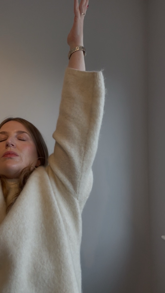 Ailbhe reaching one arm upward, eyes closed, in a soft cream jumper.
