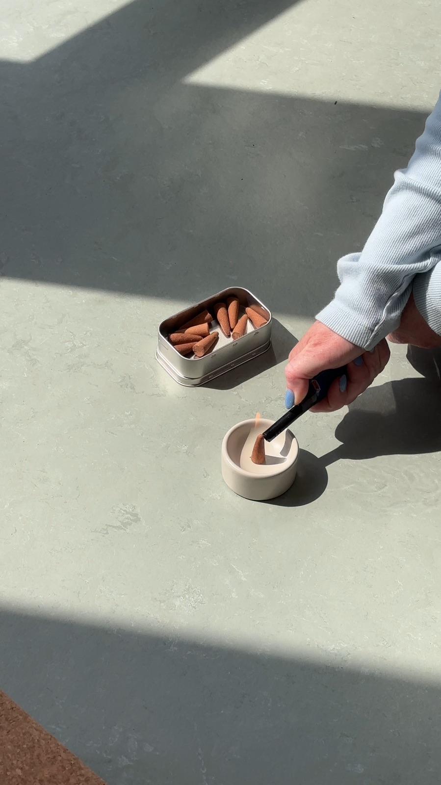 Hand lighting an incense cone beside a small tin, soft daylight.