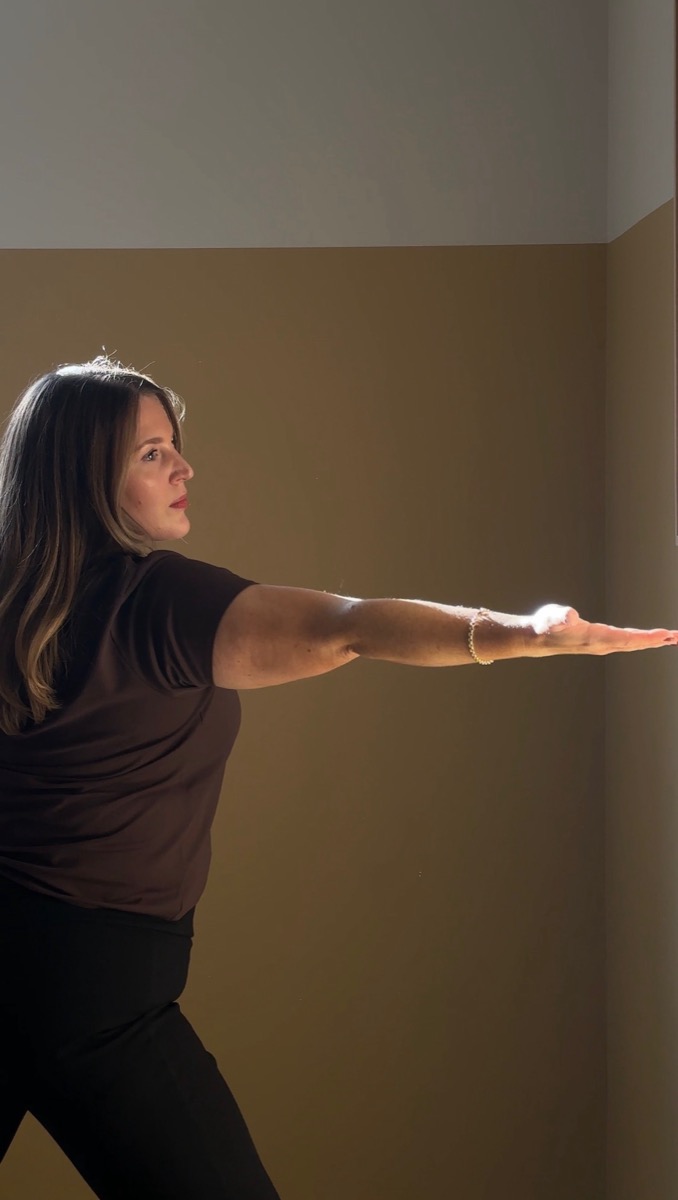 Ailbhe standing tall with one arm extended, calm and grounded in warm light.