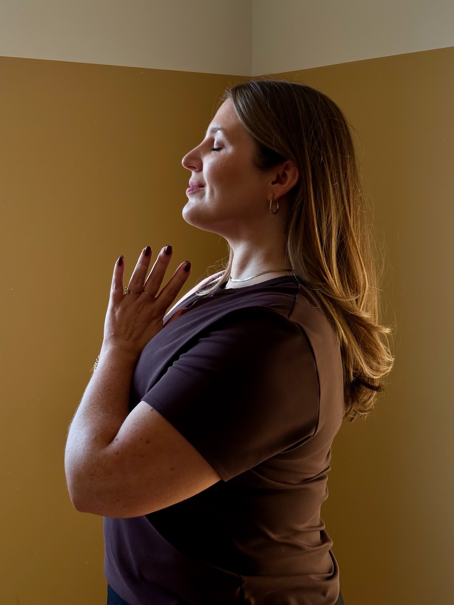 Ailbhe standing calmly with hands at heart centre, eyes closed, lit by warm afternoon light.